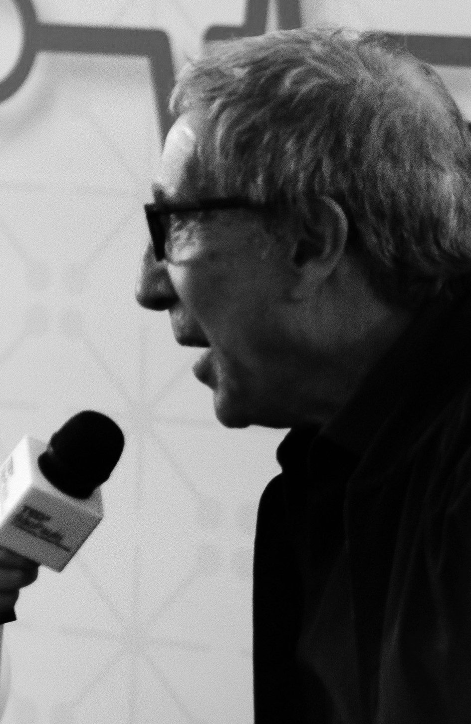 An older man with grey hair and glasses, in a dark shirt, speaks in profile towards a microphone held up to him from the left edge, in front of a pale patterned backdrop.
