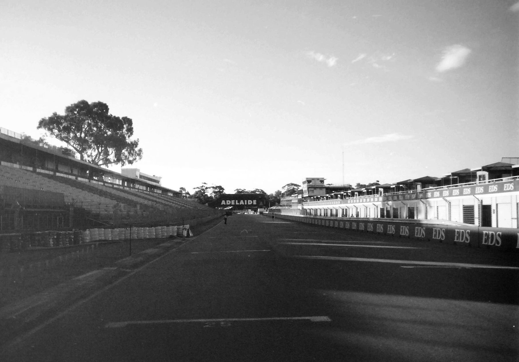 A long pit straight in low sun, empty but for two small figures far down it: a grandstand and a tall gum tree on the left, a low pit wall and two-storey pit buildings on the right, a grid number painted on the track in the foreground, and a banner bridge across the track in the distance.