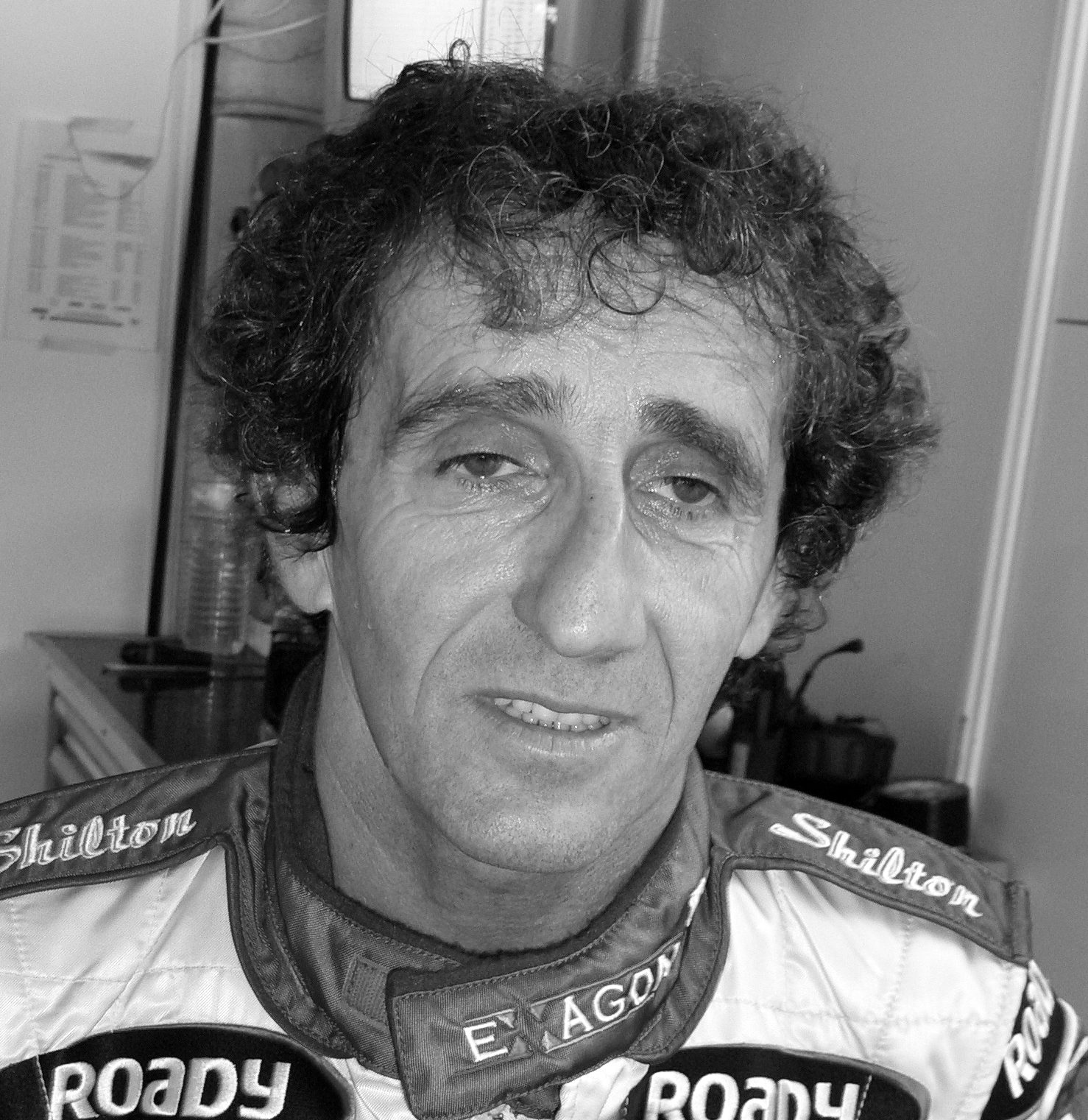 A man with a mass of dark curly hair looks straight at the camera, in a racing suit with sponsor names on the collar and chest, in a small room with a television monitor behind him.
