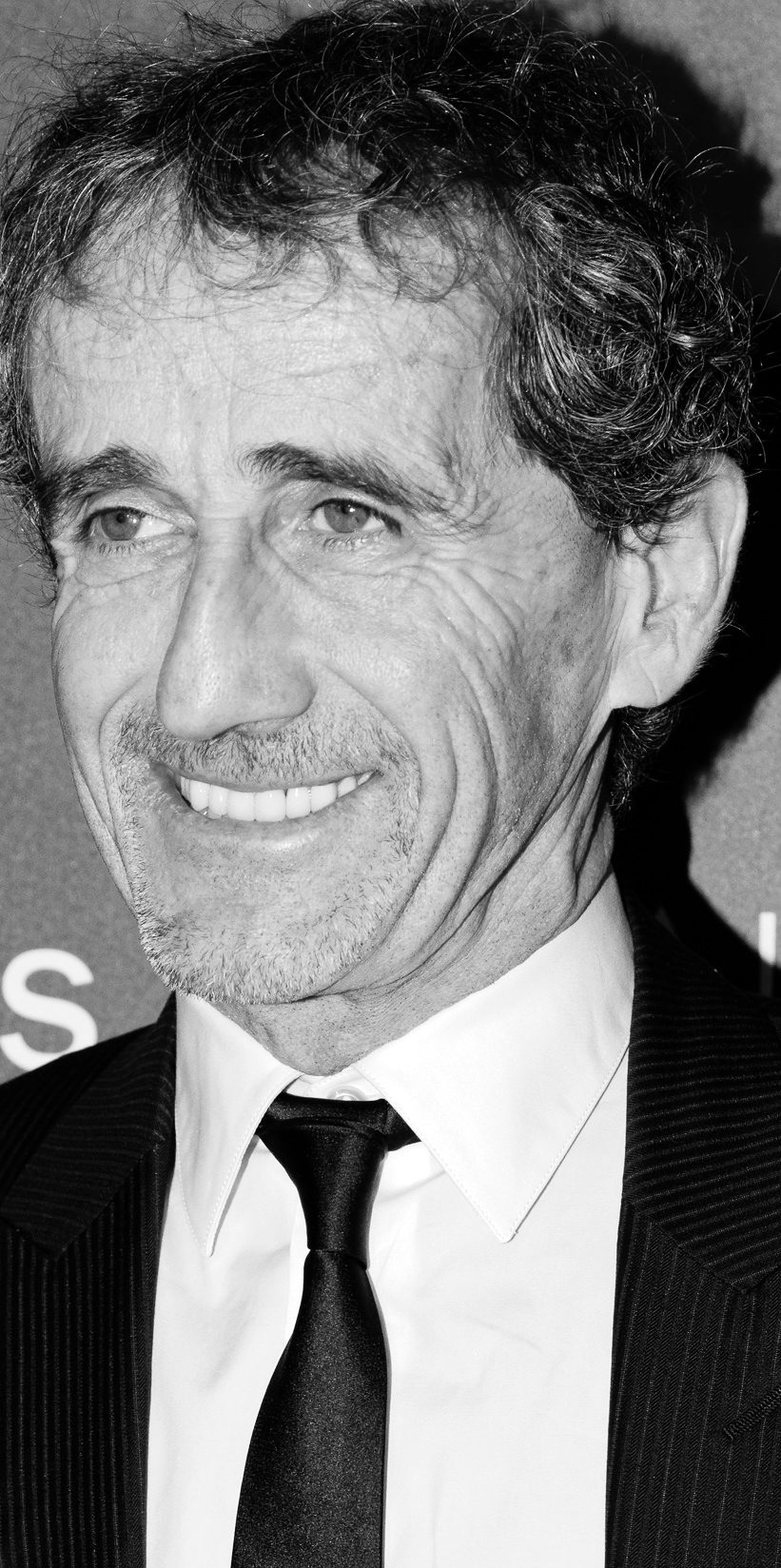 Head and shoulders of a smiling man with curly greying hair, in a dark pinstripe suit, white shirt and dark tie, against a pale backdrop with part of a large letter at its edge.