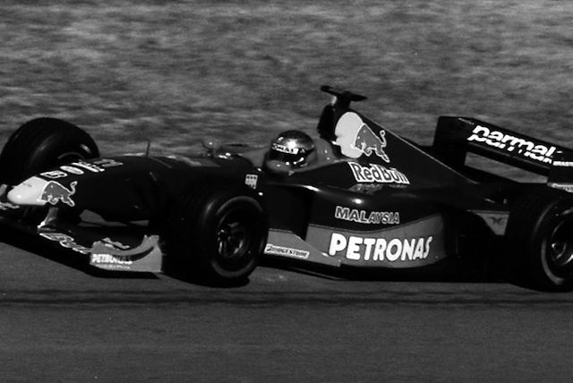 A racing car with a dark body and a charging-bull emblem on its engine cover runs along a track edged by dry grass, its driver’s helmet catching the sun.