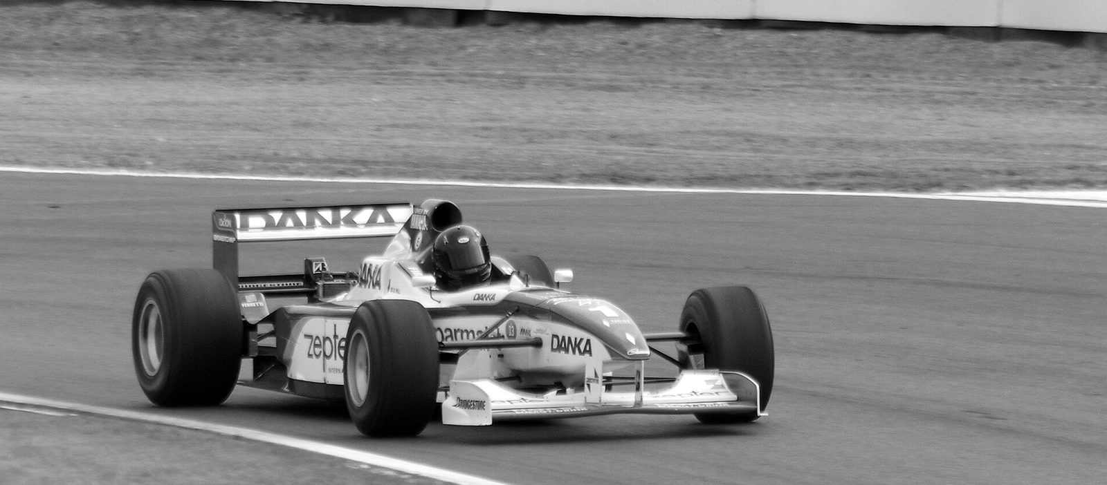 A single-seater racing car takes a left-hand bend on an otherwise empty circuit, a kerb and grass in the foreground and a dirt run-off area behind it.
