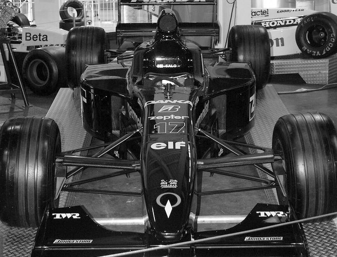 A dark racing car seen head-on on a museum plinth, the number 17 on its nose and its wide front wing filling the lower part of the frame, other exhibits behind it.
