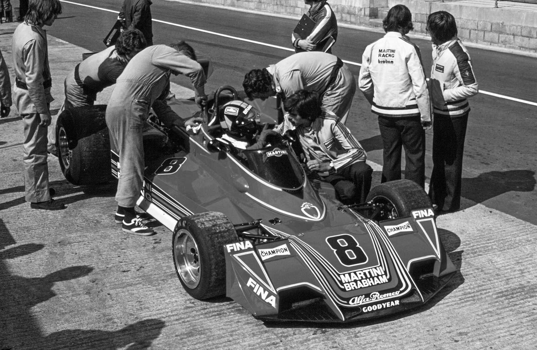 A driver in a helmet sits in a wedge-nosed racing car, number 8, in a pit lane; three mechanics in overalls lean over the car, a man in a team jacket crouches by the cockpit, and others in overalls and team jackets stand round it.