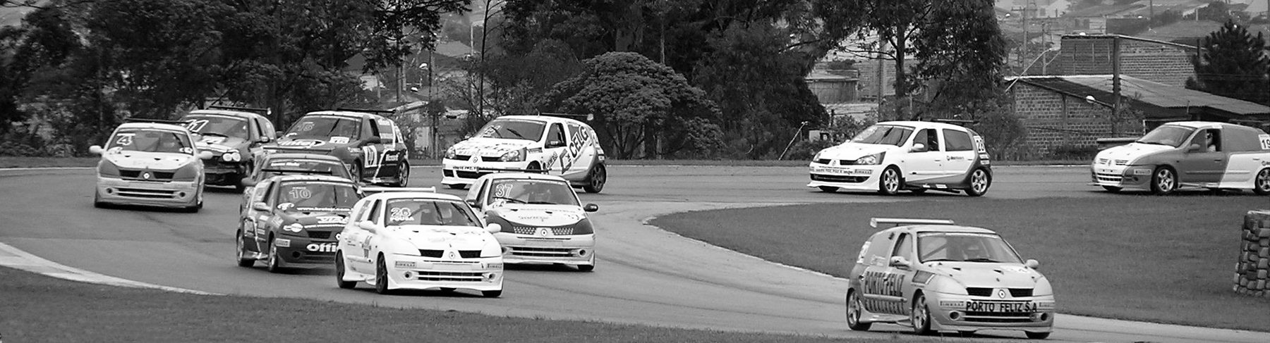 A pack of small hatchback racing cars rounds a left-hand bend across a grassy infield, with trees and houses beyond the circuit.
