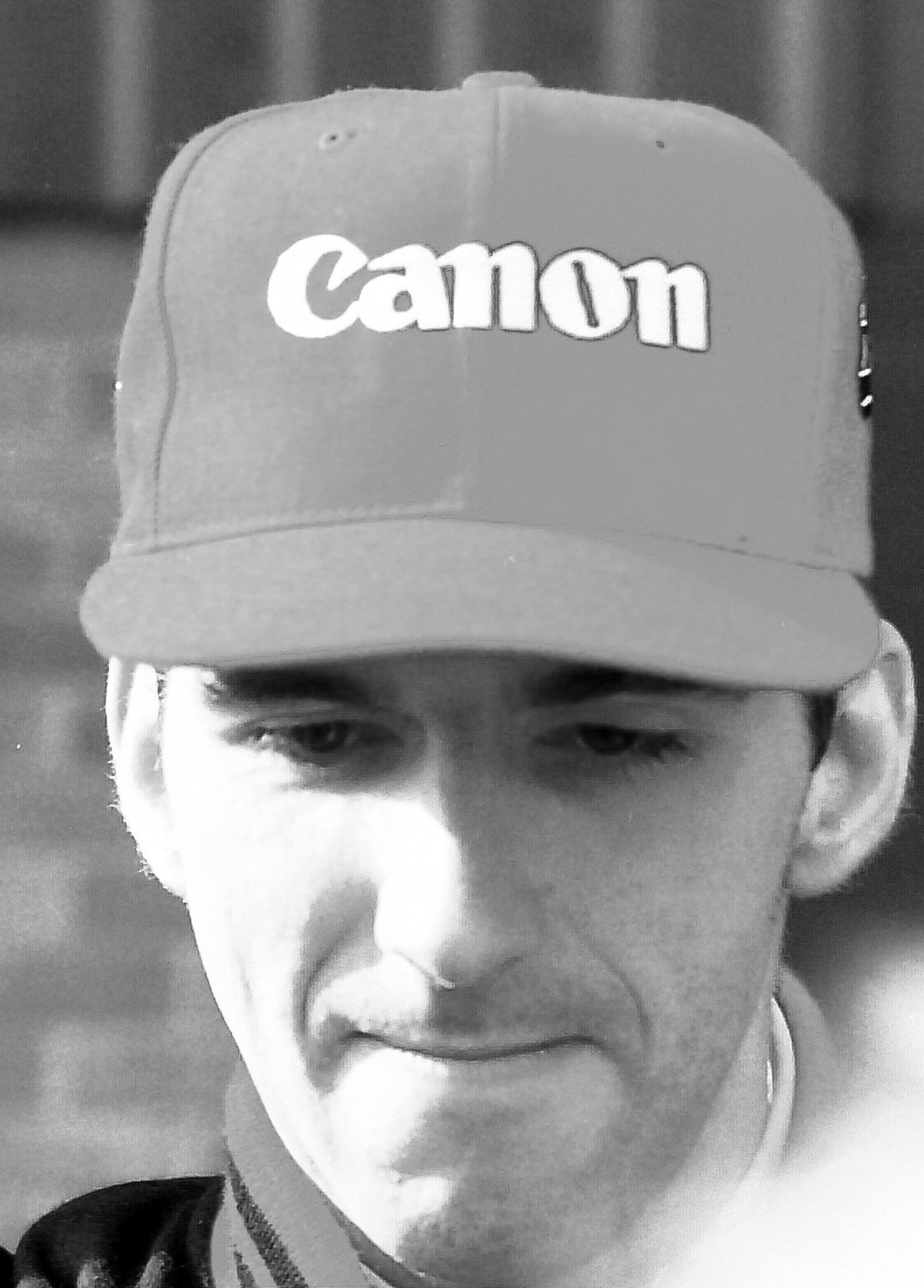 Close portrait of a young man in a peaked cap bearing a sponsor’s name, his eyes lowered, in front of a brick wall.
