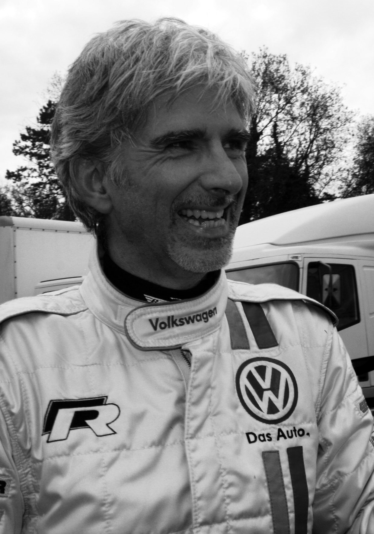 A grey-haired man with a short beard laughs as he talks, in a pale racing suit with sponsor badges, with lorries and trees behind him.