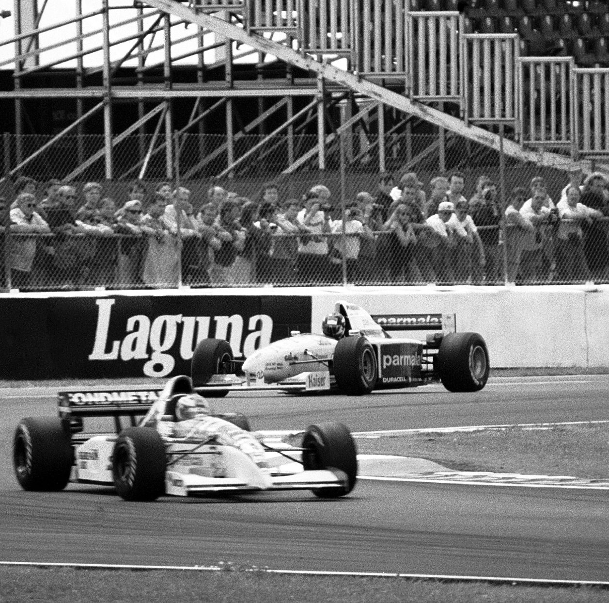 A white racing car leads another single-seater, its bodywork covered in sponsor names, out of a corner; behind them spectators line a catch fence below the scaffolding of a temporary grandstand, above a long advertising board.