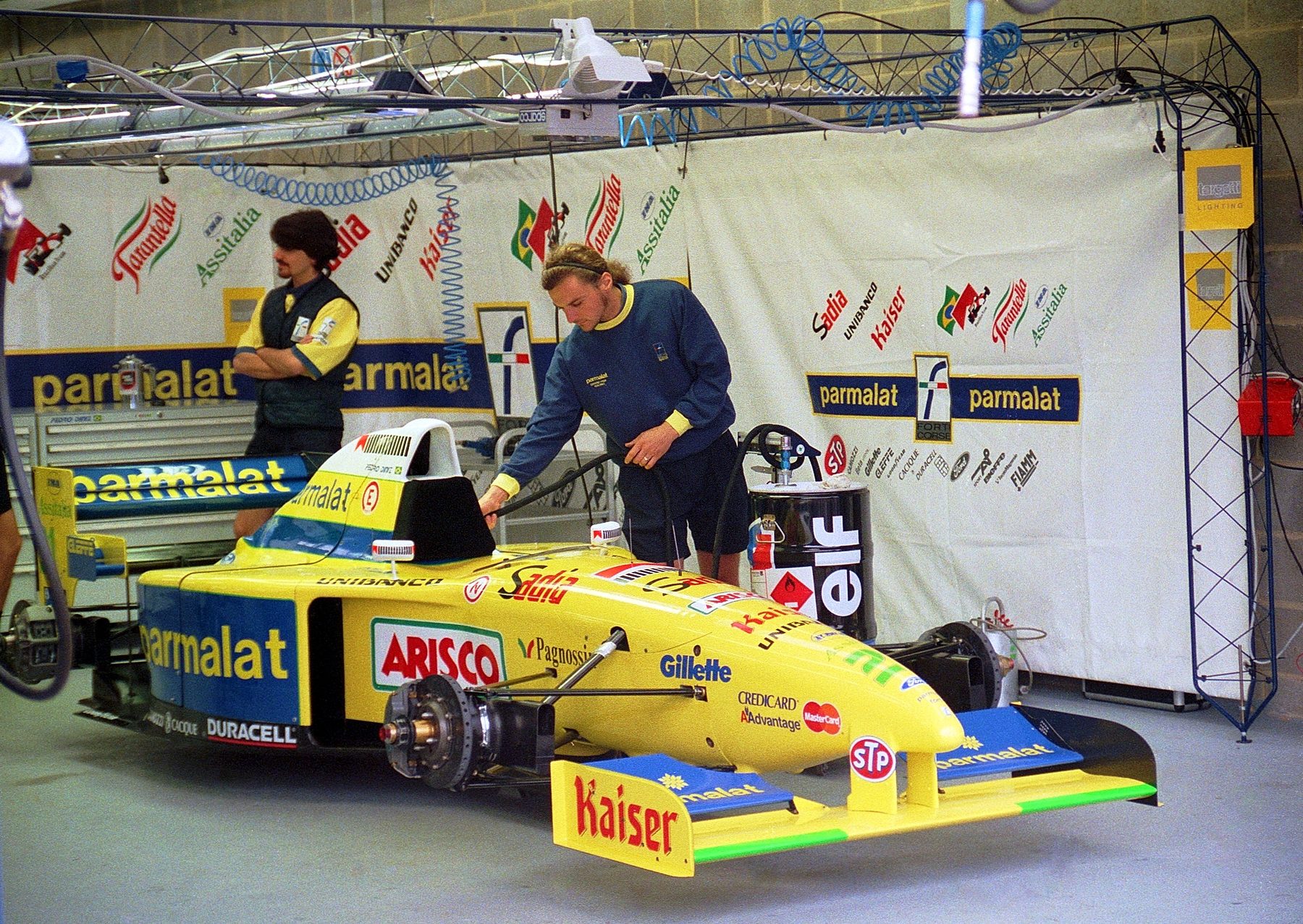 A single-seater racing car stands with its wheels off in a pit garage, its bodywork covered in sponsor names; one mechanic leans into the cockpit while another stands behind with folded arms, in front of a backdrop printed with more sponsor logos.