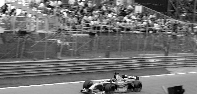 A single-seater racing car, blurred with speed, passes along a straight in front of a grass verge, a guardrail and a crowded grandstand.