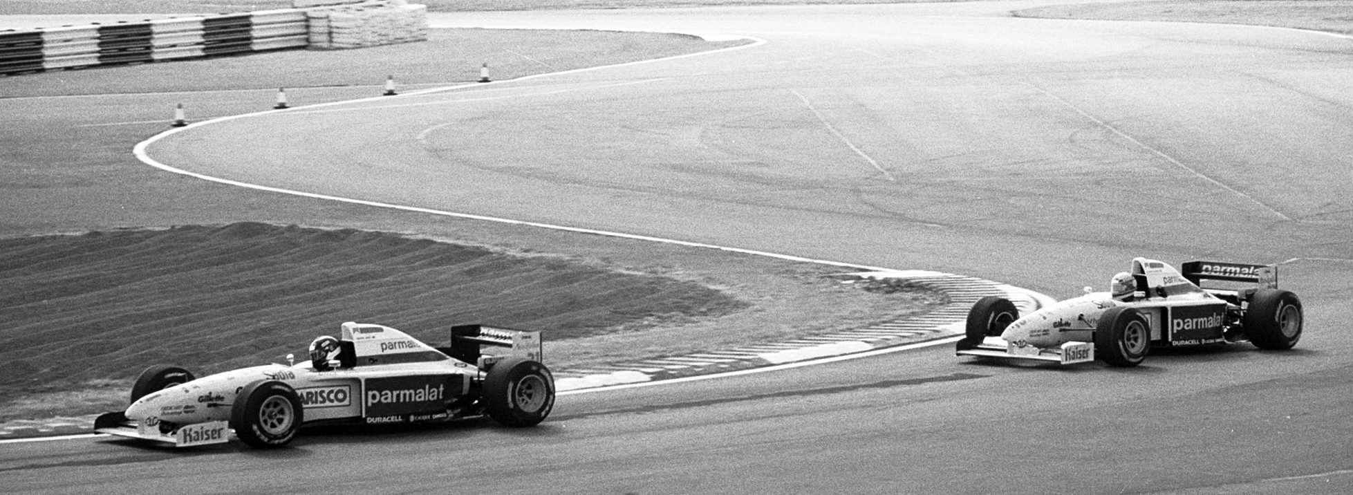 Two single-seater racing cars in the same sponsor-covered livery round a sweeping curve of empty track, one some lengths behind the other; beyond them lie a raked gravel trap, grass and run-off, with a tyre barrier at the far edge.