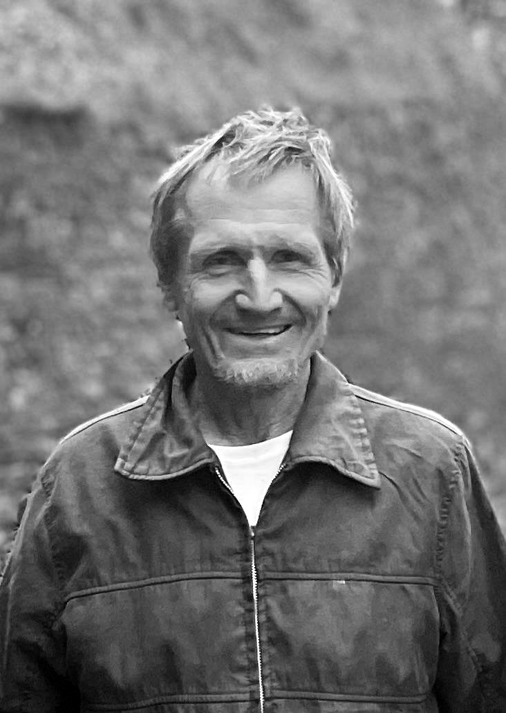 Head-and-shoulders portrait of a smiling man with tousled fair hair and a short beard, in a zipped jacket over a T-shirt, against a mossy stone wall.