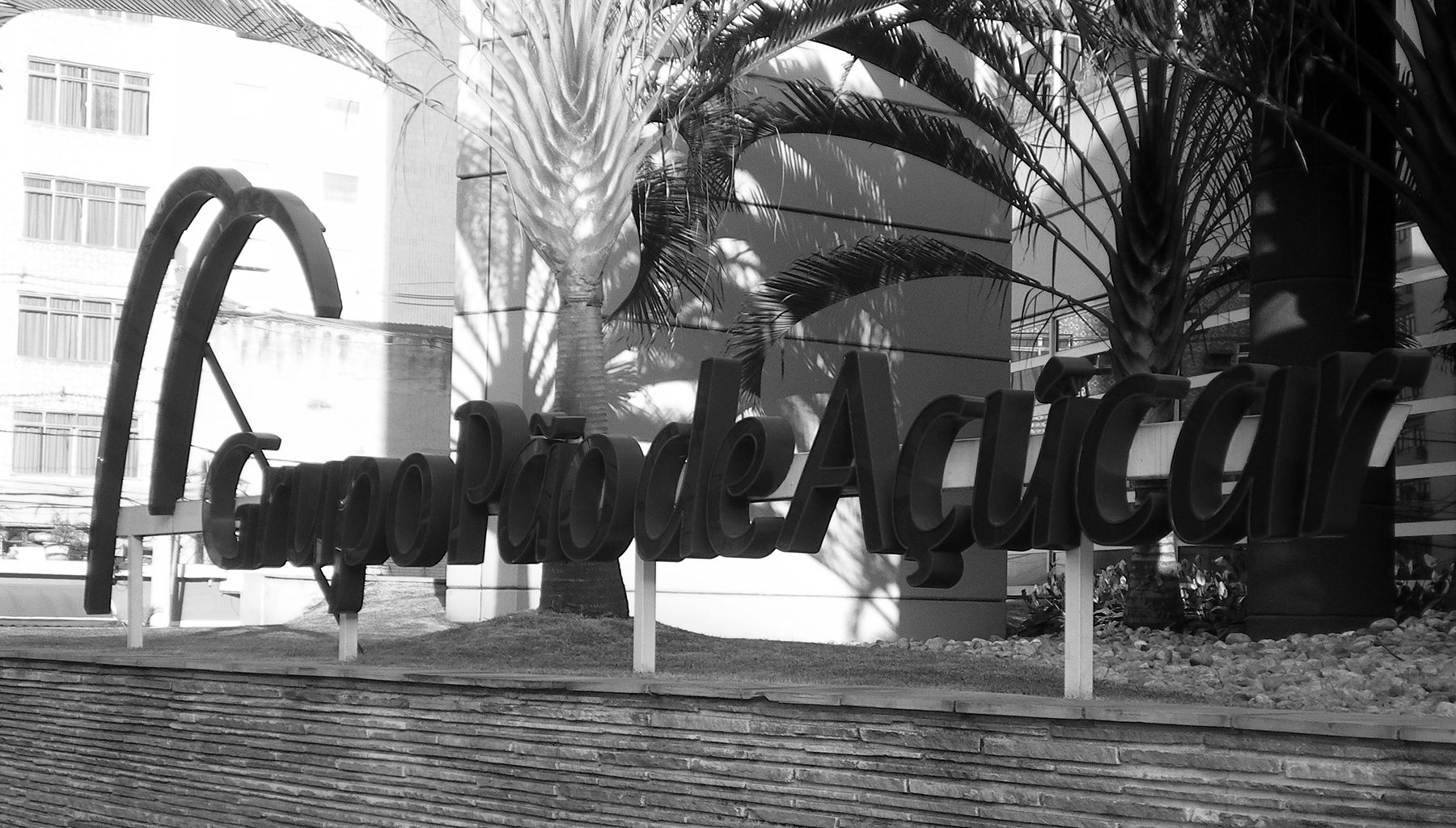 Large script letters reading Grupo Pão de Açúcar, beside a logo of two arches, stand on posts along the top of a low stone wall under palm trees, with office buildings behind.