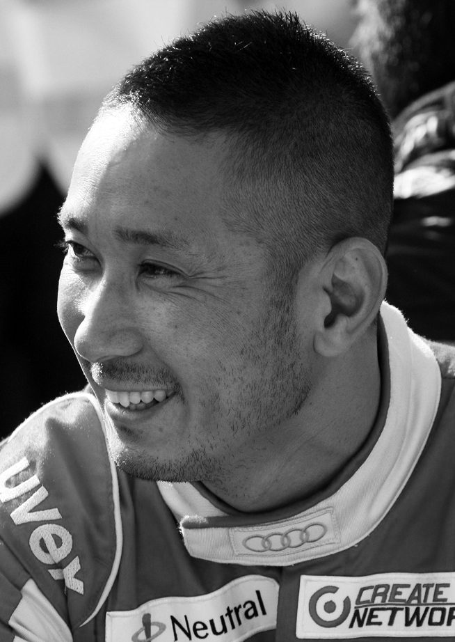 A smiling man with close-cropped hair looks off to the left, in a racing suit covered in sponsor patches, with people blurred behind him.