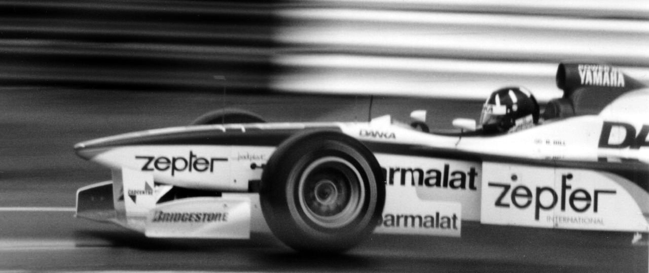 A racing car photographed at speed against a streaked, blurred background, its pale bodywork carrying sponsor names; the driver’s dark helmet shows above the cockpit.