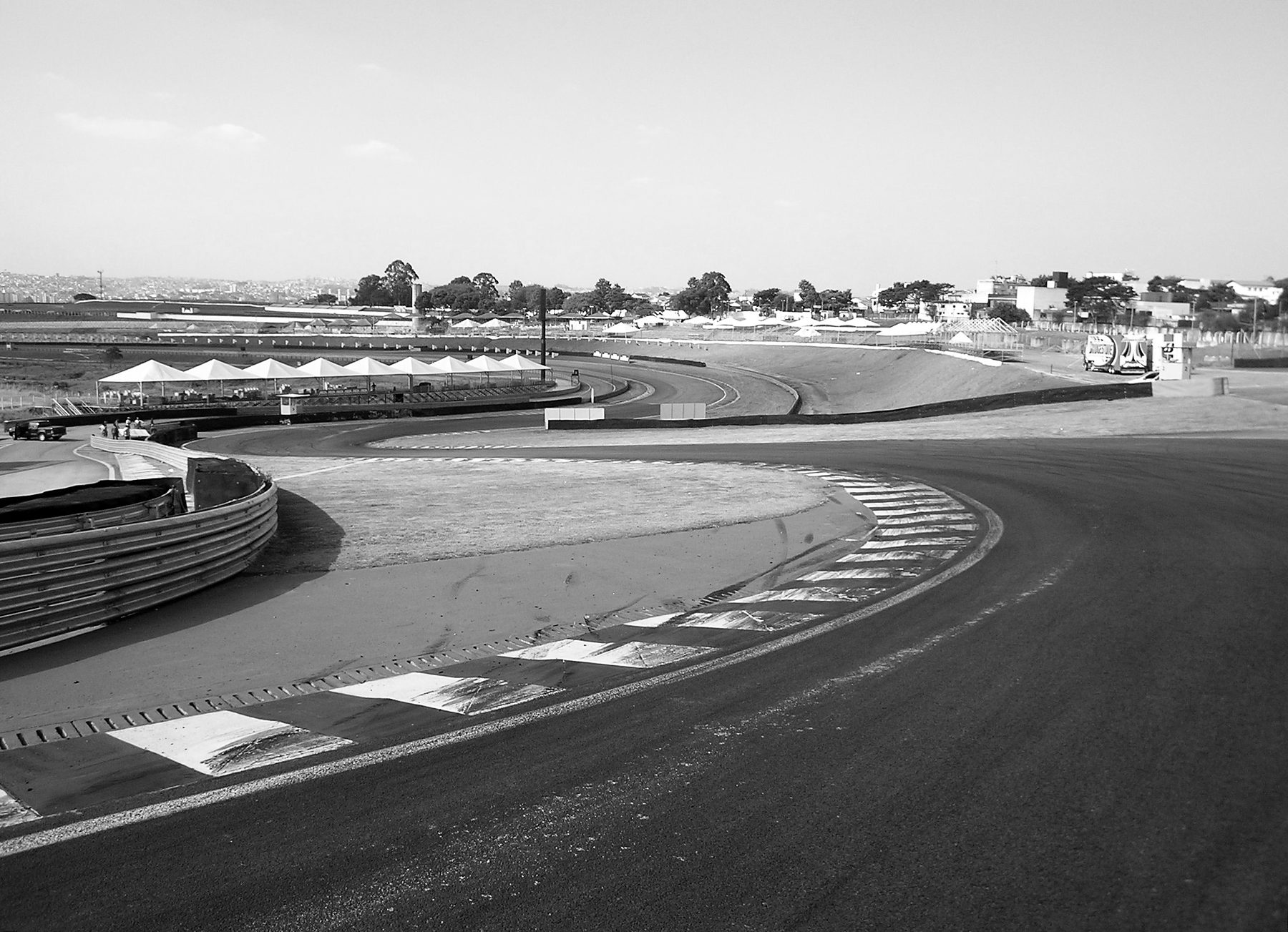 An empty racing circuit seen from beside the track: a sweeping left-hand bend with striped kerbs and painted run-off in the foreground, the track curving away past white tents and grandstands towards the houses of the city.