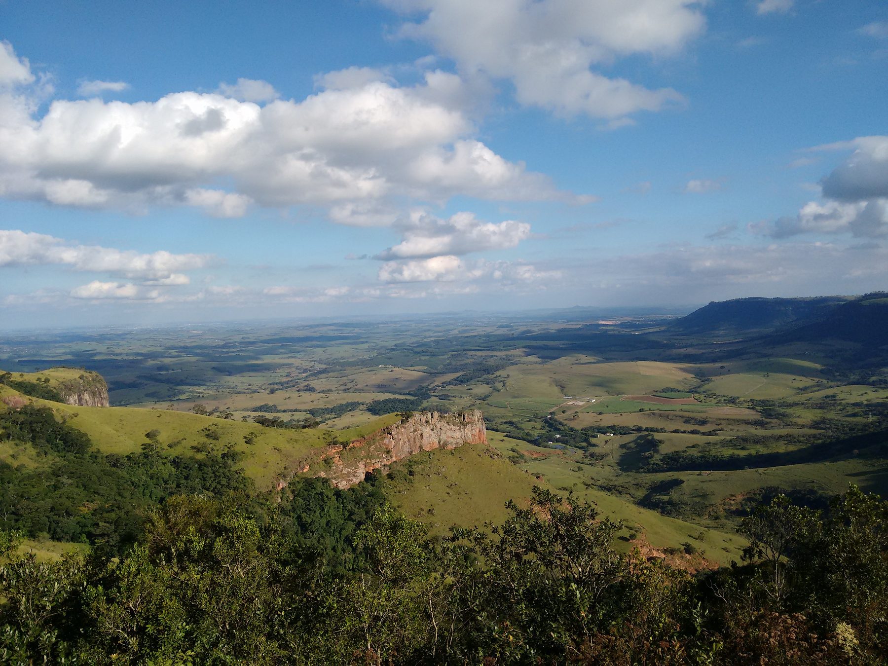 From a wooded hilltop, a pale sandstone cliff with reddish faces juts out over rolling green farmland and patches of dark forest that stretch to a hazy horizon, under a blue sky and heaped white clouds, with a long escarpment rising on the right.