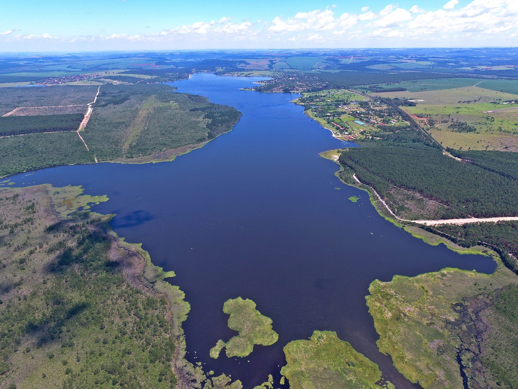 An aerial view along a long, winding reservoir: marshy islands and scrub in the foreground, a strip of houses along the far right shore, and plantations and patchwork fields beyond it to the horizon.