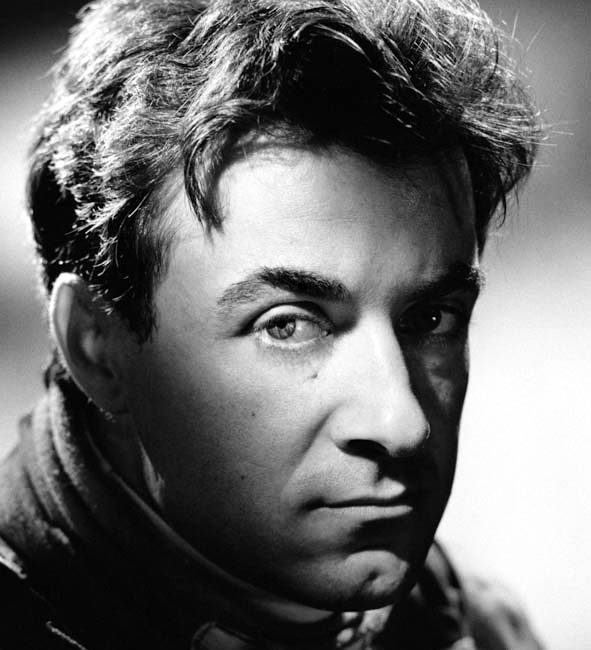 A studio portrait of a man with tousled dark hair, lit hard from one side, looking into the camera over the high collar of a racing suit.