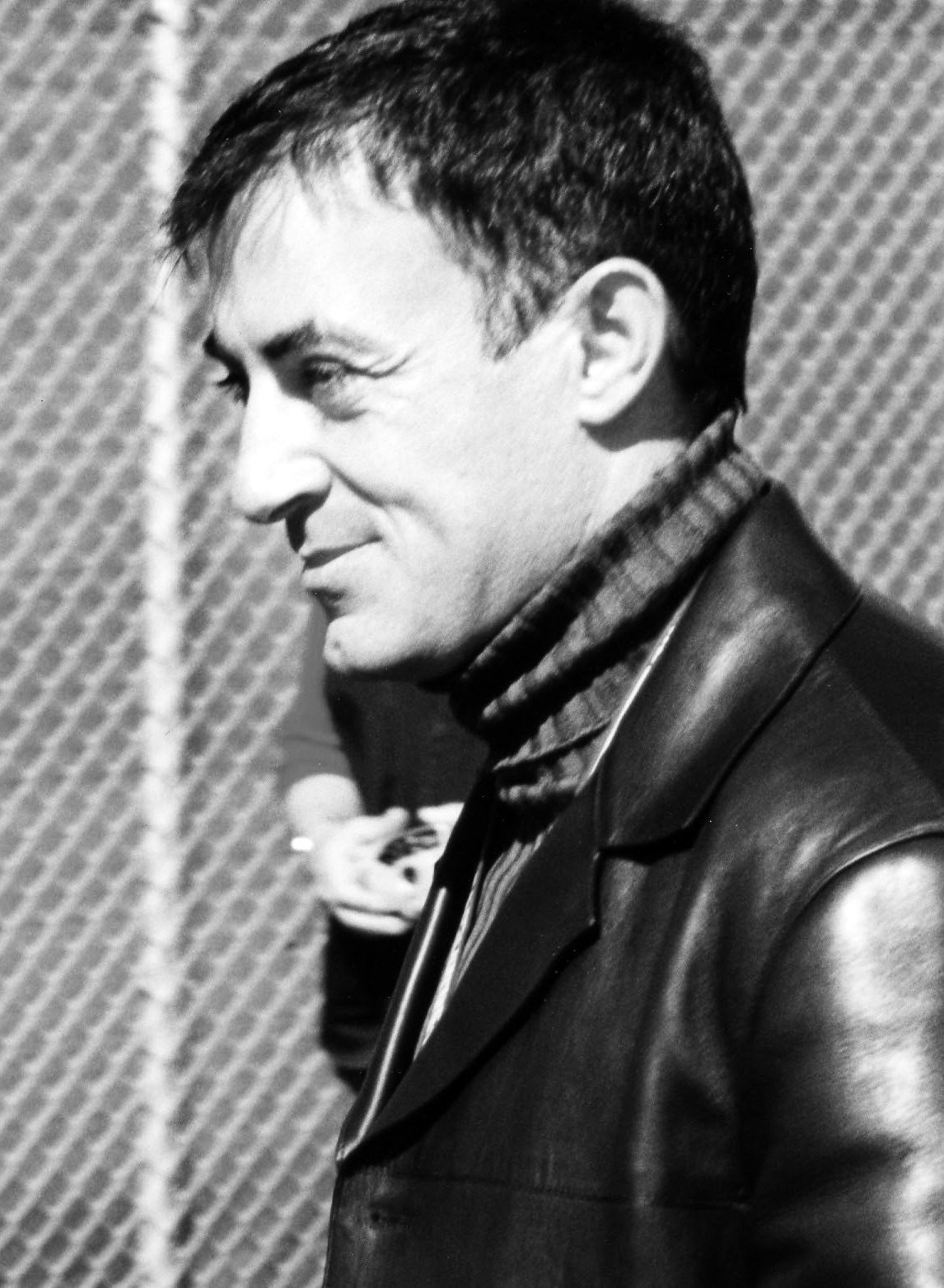 A dark-haired man in a leather jacket and a knitted scarf, smiling, seen in profile against a wire-mesh fence, with someone partly visible behind him.