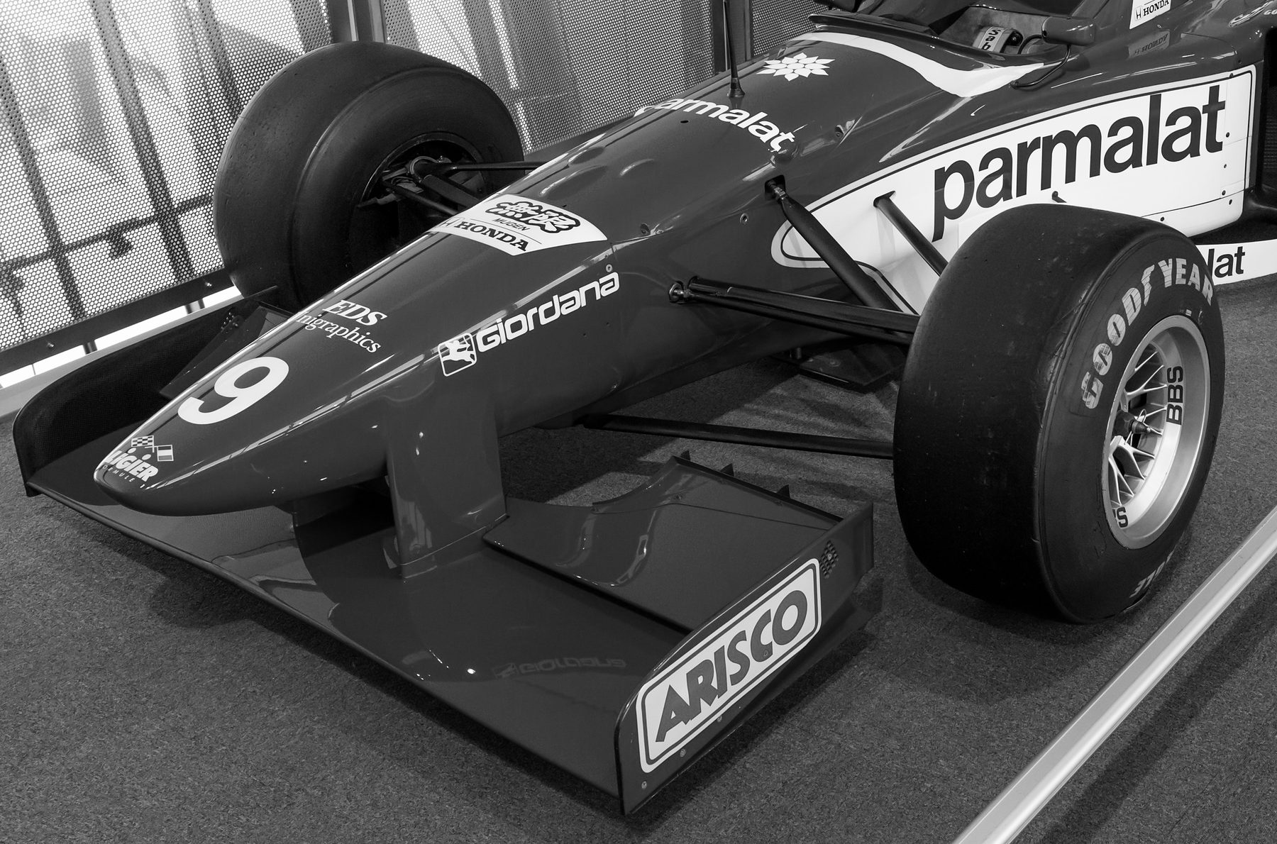 The nose, front wing and front wheels of a racing car on a museum floor: a large number 9 on the nose, sponsor names along the body and on the wing endplate, and a railing behind.