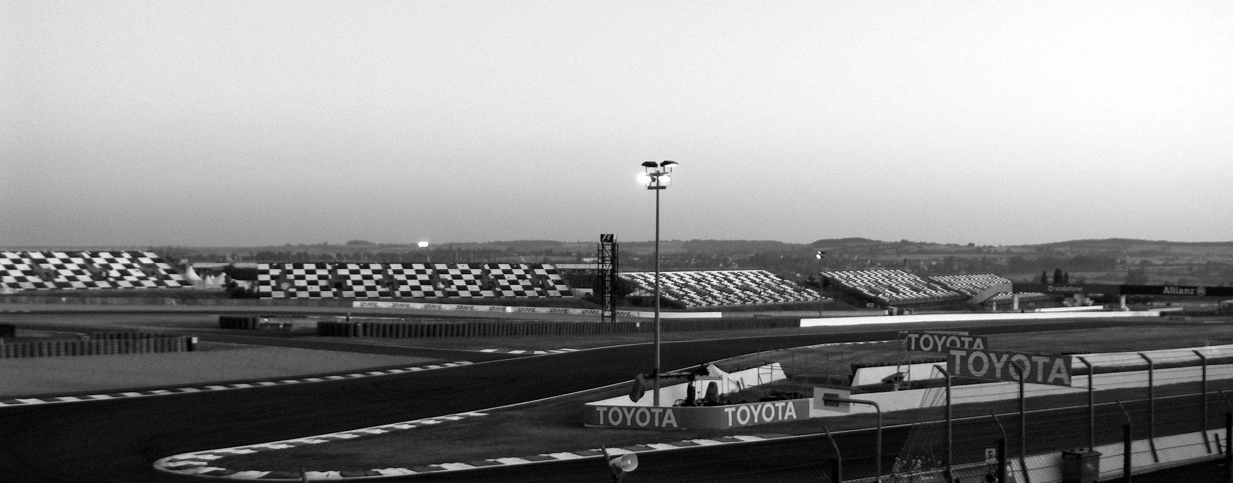 A circuit at dawn: a tight hairpin edged with striped kerbs, advertising boards and a floodlight on a tall mast, and grandstands with chequered seating along the horizon under a pale sky.