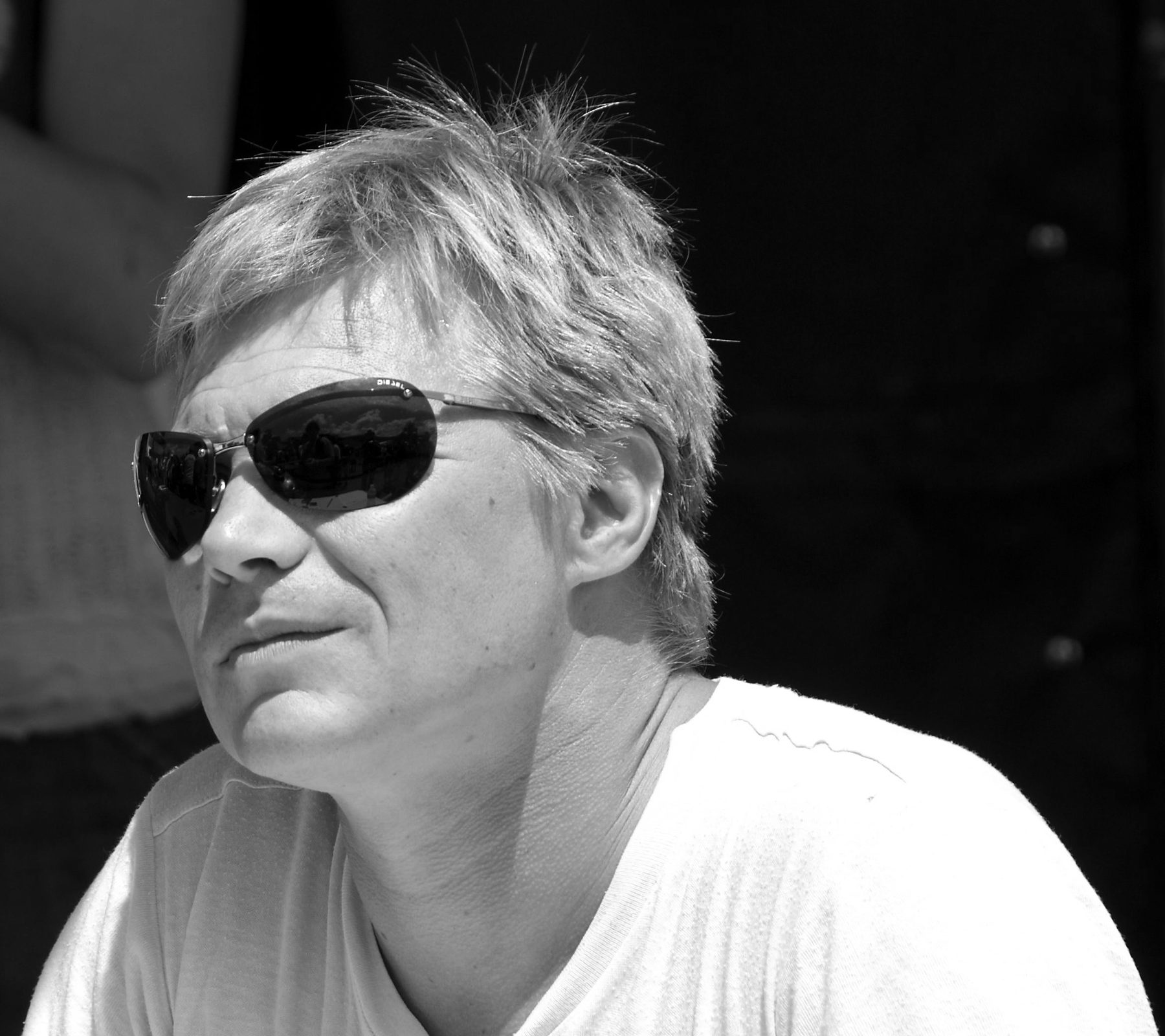Head-and-shoulders portrait of a man with tousled fair hair and wraparound sunglasses, in a pale T-shirt, looking off to the left against a dark background.