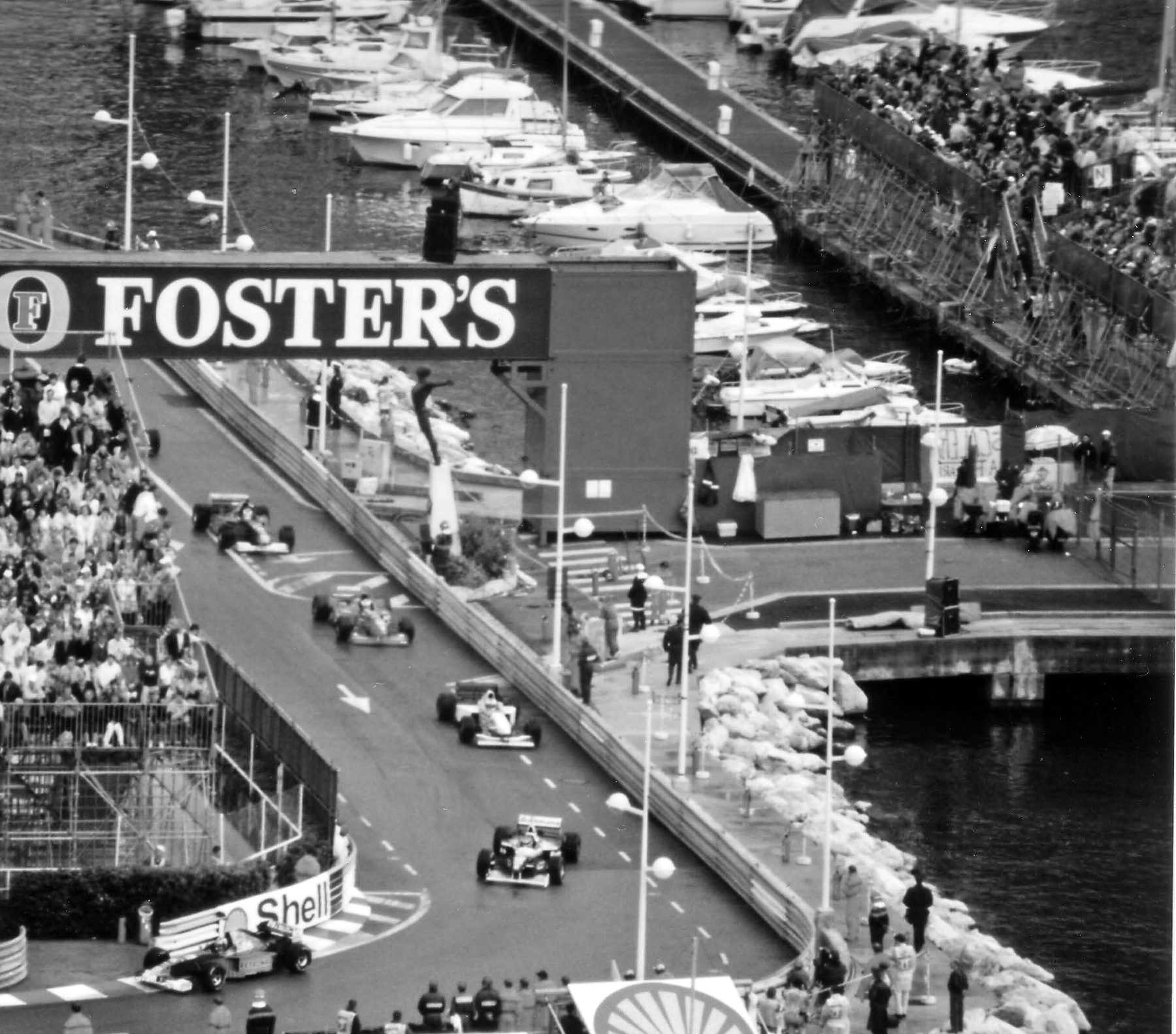 Seen from high above, racing cars run in single file along a harbourside road between packed temporary grandstands and a quay lined with moored yachts, below a footbridge carrying a large advertising banner.