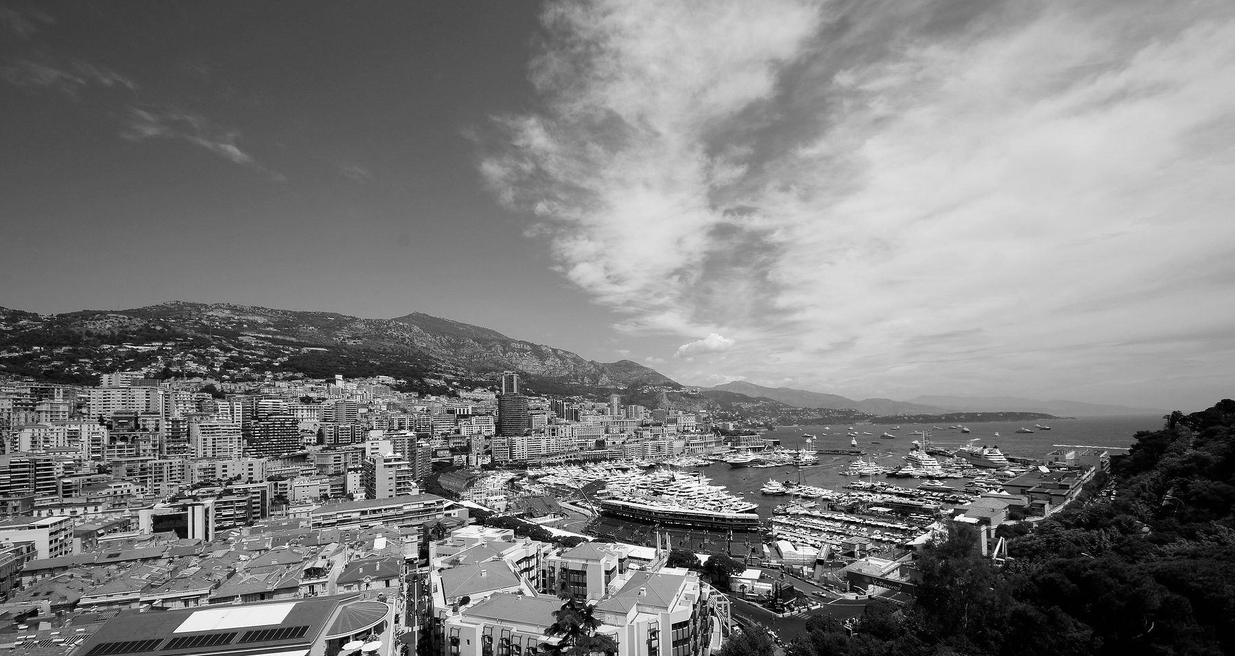 A hilltop view over a crowded harbour: tiled roofs and apartment blocks in the foreground, rows of moored yachts and temporary grandstands along the quays, and the town climbing a mountainside under a wide sky.