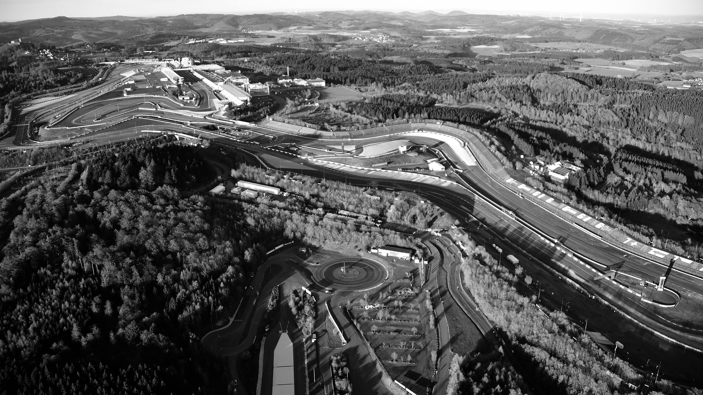 An aerial view in low evening light over a racing circuit set among forested hills: the pit straight, grandstands and pit buildings in the middle distance, a roundabout and service roads in the foreground, and farmland rolling to the horizon.