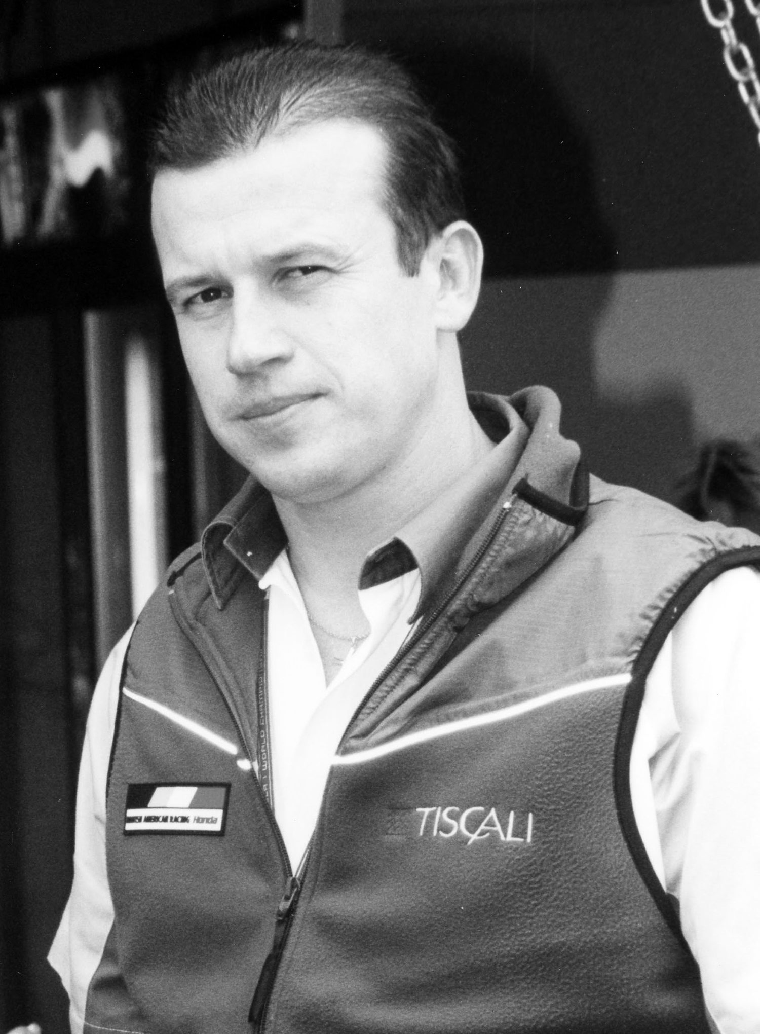 A man with short, combed-back hair and a wary half-smile, in a fleece gilet with a team badge over an open-necked shirt, stands in front of a dark doorway.