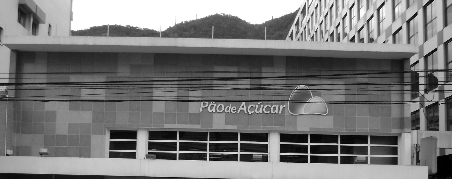 The upper façade of a supermarket, clad in square tiles of two tones, with the name Pão de Açúcar and a logo of two rounded hills above a row of windows, and a wooded hill behind the building.