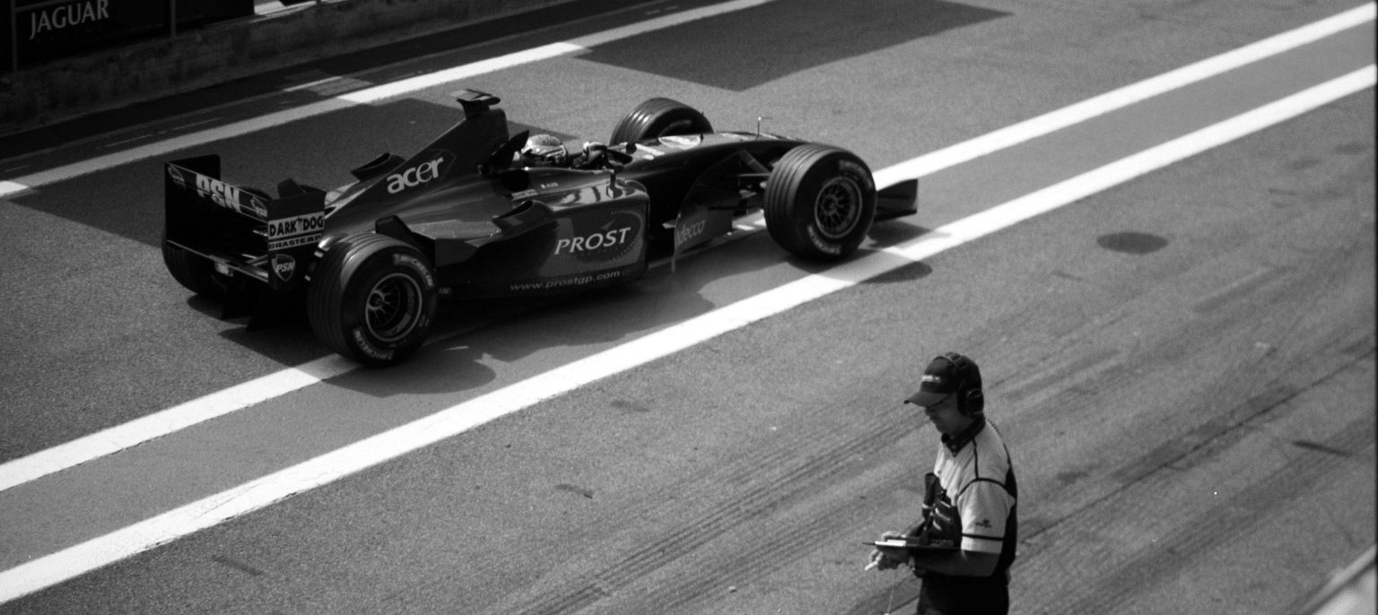 A racing car pulls away along a pit lane marked with a broad painted stripe, while a team member in a cap and headphones looks down at a clipboard in the foreground.