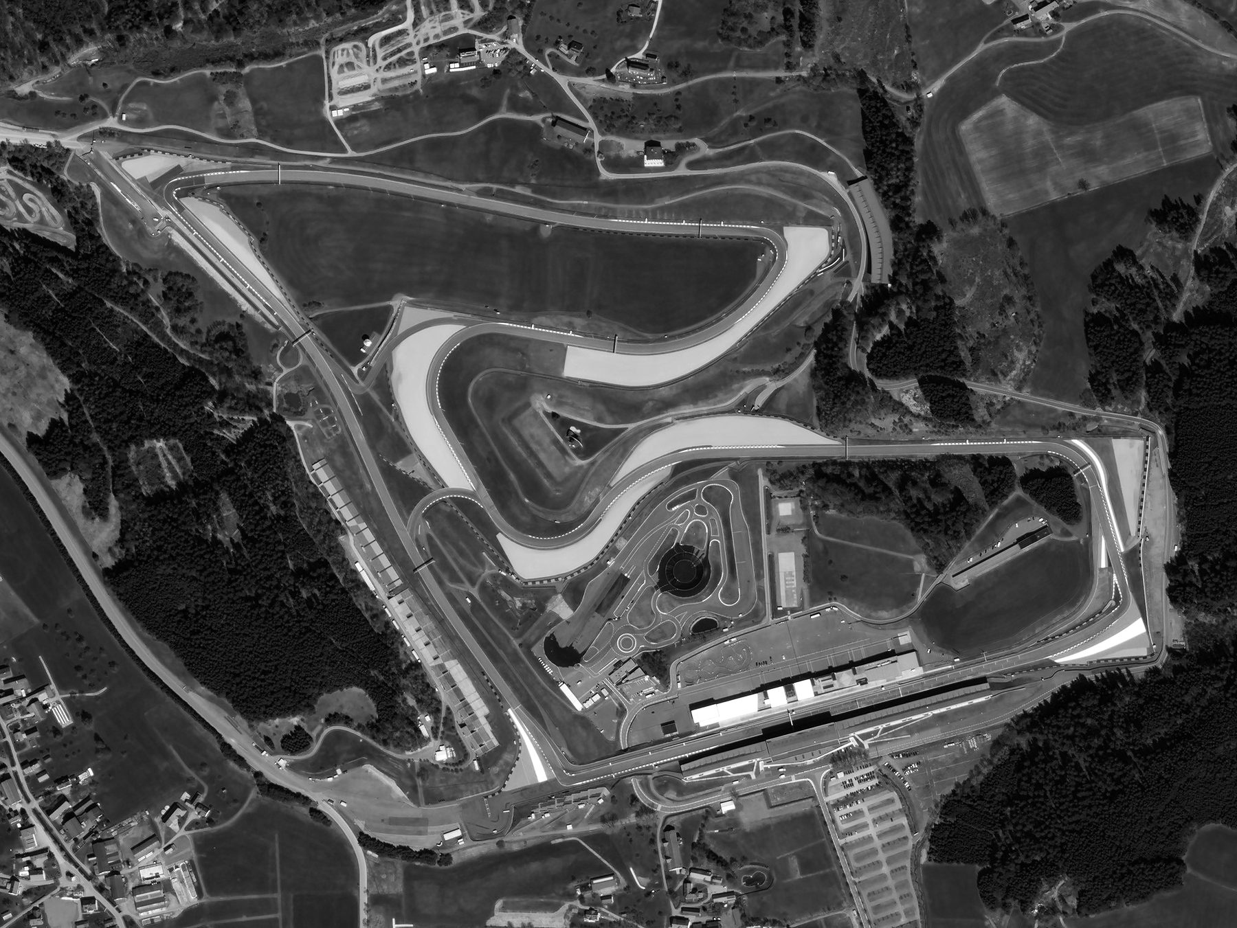 A satellite view of a racing circuit among fields and forest: the track loops round a sloping infield, with long pit buildings and grandstands beside the straight at the bottom and a village at the lower left.