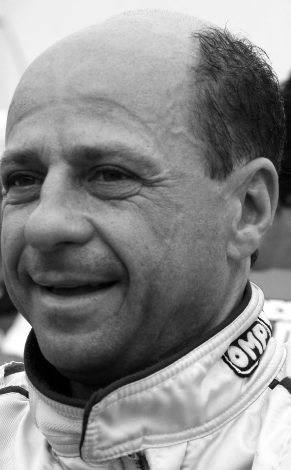 A balding, smiling man in a pale racing suit looks off to the left, with other people blurred behind him.