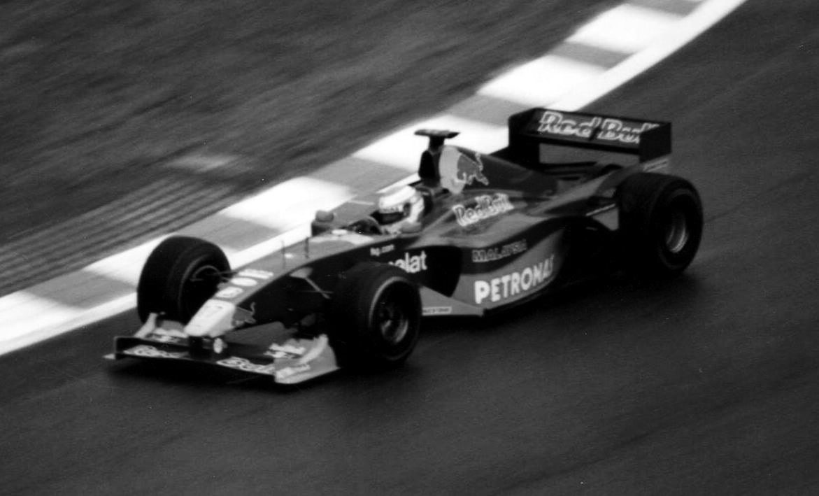 A racing car runs on a wet, dark track beside a striped kerb, the grass behind it blurred by the camera’s pan; the driver wears a light helmet.