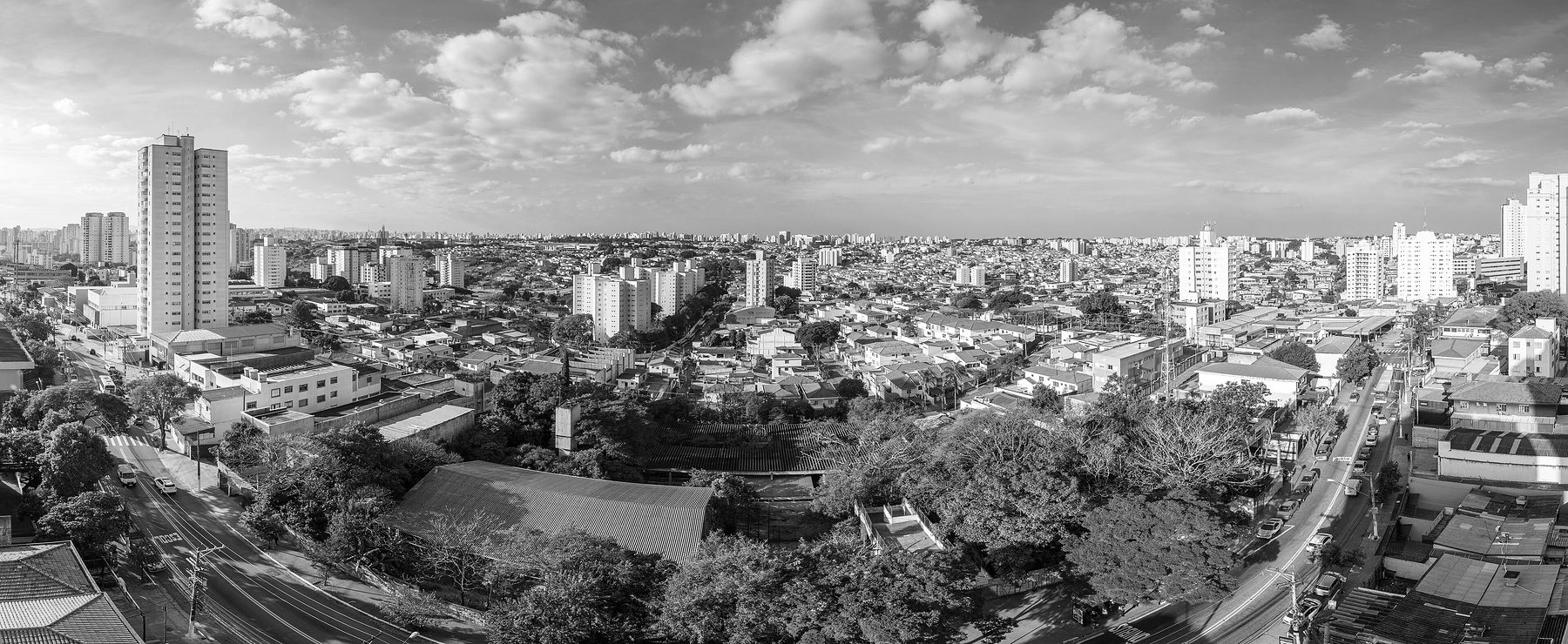 A wide view over a city: low houses with tiled roofs and scattered tower blocks stretching to the horizon under a sky of broken cloud, a busy avenue running along the left edge.