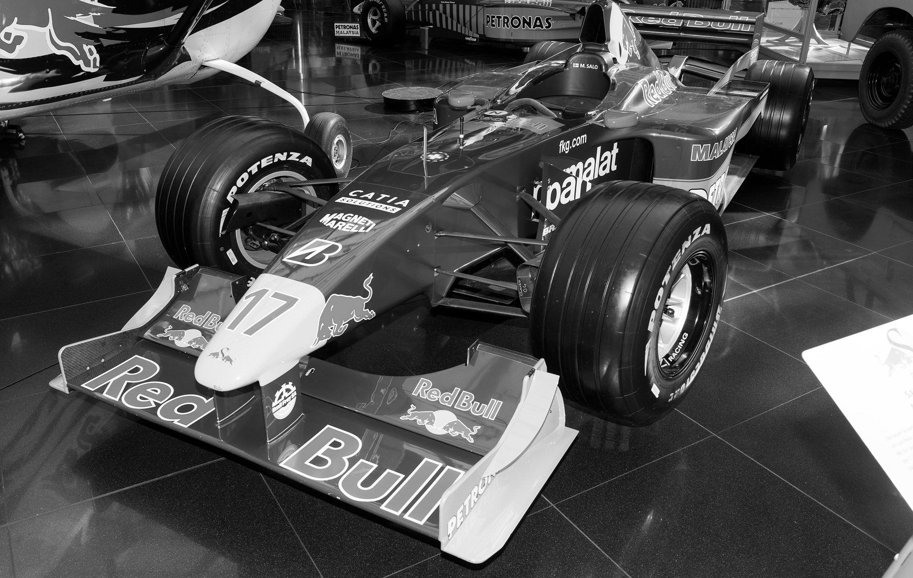 A racing car, number 17, seen from the front left on a polished floor, sponsor names across its nose and front wing, the underside of a light aircraft at the top left and another car behind.