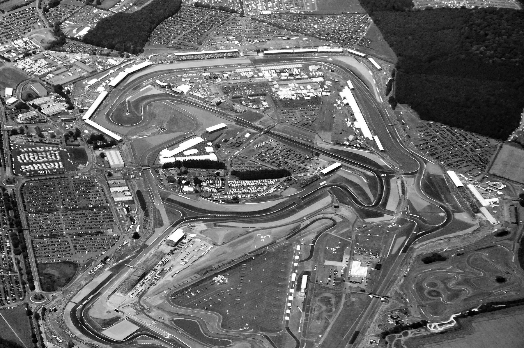An aerial view of a racing circuit in open countryside, the track looping among grandstands, hospitality tents and vast car parks full of cars, with woodland on the right.