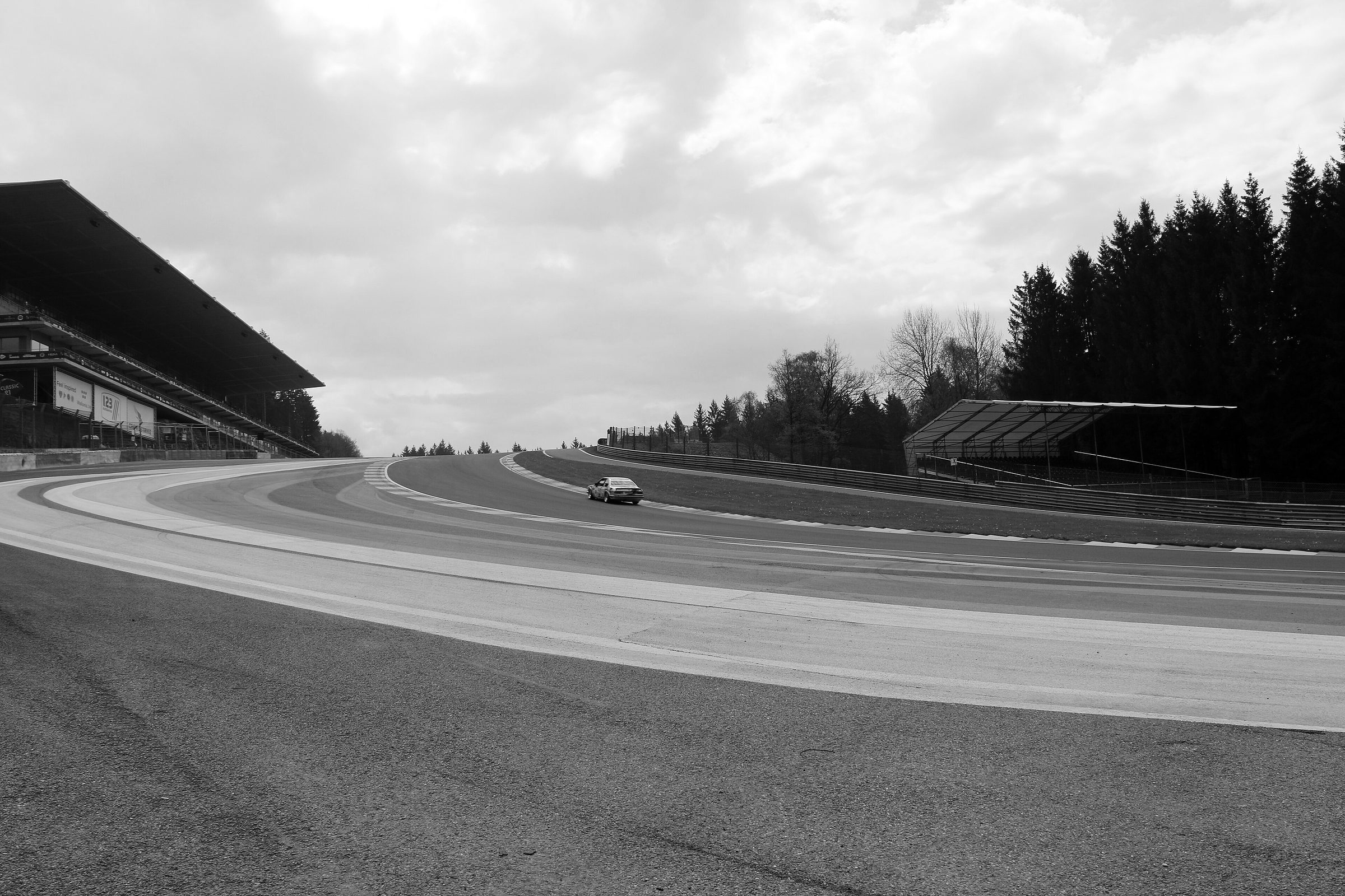 Seen from track level, a circuit climbs steeply uphill through a left-right bend; broad painted kerbs run across the foreground, a covered grandstand stands on the left, pine forest on the right, and a single saloon car heads up the slope.