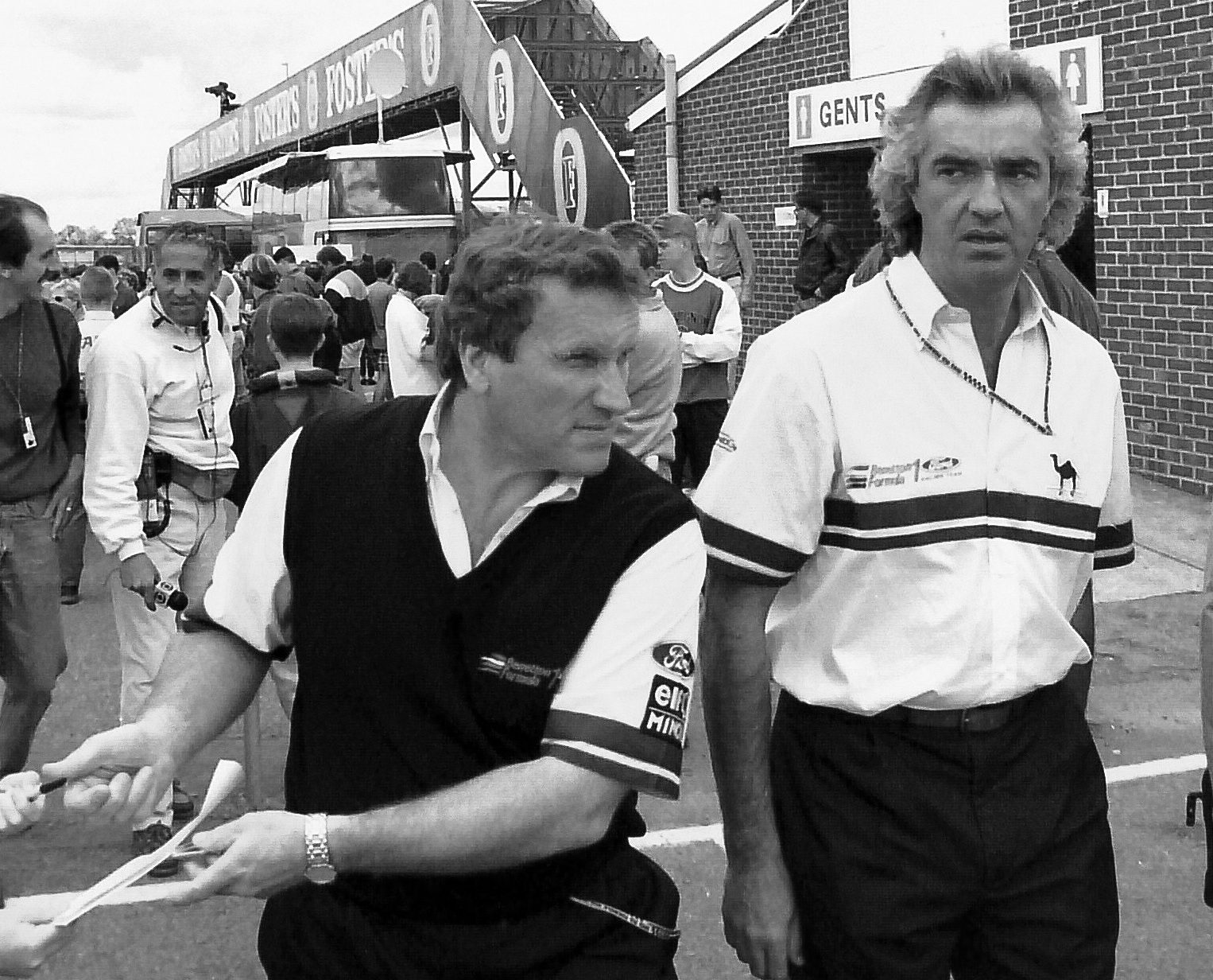 Two men in team shirts walk through a crowded paddock. The one in front, in a dark sleeveless jumper, holds out a pen and paper to a child’s outstretched hand, while a taller man with swept-back grey hair looks towards the camera; a footbridge and a brick building stand behind them.